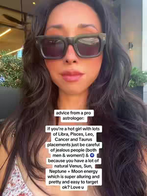 A selfie featuring a woman with long dark hair, wearing large, dark sunglasses. She has visible makeup and is positioned against an out-of-focus outdoor background, likely a patio or similar setting. Text overlays provides astrological advice. The overall composition is a close-up portrait with a focus on the face.