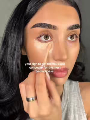 A close-up of a woman with dark hair applying concealer under her eye with an applicator. The image is well-lit, with a focus on the woman's face and the makeup application. The background is blurred, keeping the focus on the foreground. A text overlay on the image suggests it promotes a specific concealer.