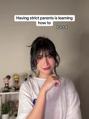 A woman with dark hair and bangs smiles at the camera. She wears a white t-shirt and has red lipstick on. Behind her is a shelf with several small character figurines and a potted plant.