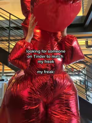A person wearing a full-body, red, inflatable costume that resembles a large heart or balloon structure, standing in what appears to be an office setting with stairs in the background. The person's hands are raised, and text overlays appear on the image.