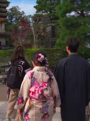 The cover image shows three people walking along a path, with two of them in traditional Japanese attire. The foreground features a person in a kimono with a large pink bow, and a flower in the hair. The person in a black jacket and another in dark clothing are also included, creating a sense of movement. The background includes greenery and architectural elements, suggesting a temple or historical site.
