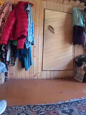 The image shows a close-up of a room. The room is constructed with wooden planks, forming the walls and likely the floor and door. On the left side, various clothes, including jackets, hang on hooks. In the center, a wooden door with a handle is visible, and on the right side, more clothing or items hang. The floor is a reddish-brown color transitioning to a lighter color, possibly a rug visible in the foreground.