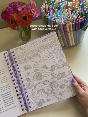 The cover image features an open coloring book with a sea-themed illustration. A hand, with blue painted nails, holds the book. The book is open to a spiral-bound page, showing an underwater scene with seashells and aquatic plants. Beside the book are a clear vase of pink and orange flowers and a clear container filled with colorful markers. The background is a white surface.
