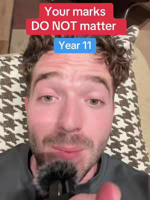 A man with stubble looks at the camera from a slightly top-down angle and holds a microphone. Text overlays state "Your marks DO NOT matter" in a red box and a blue box displaying "Year 11". The man is wearing a dark gray shirt and the background behind him is a patterned design.