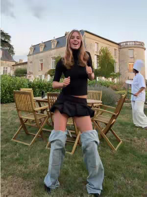 A young woman stands in front of a large building with a castle-like appearance. She is wearing a black top, a short black skirt, and denim leg warmers. The background includes a manicured lawn, wooden chairs at a table, and another person in a white outfit. The overall lighting suggests an outdoor setting with natural light.