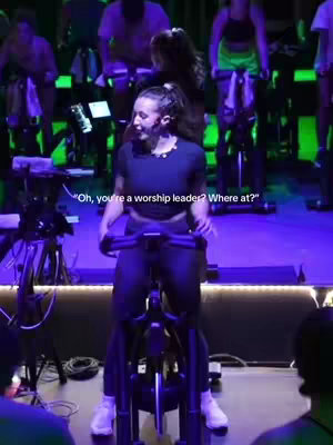 Shot of a woman on a stationary bike in a cycling studio. She is positioned in the center, looking towards the camera. There are other people on bikes in the background with various studio lights.