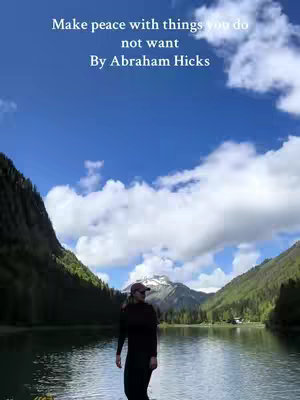 A scenic view featuring a serene lake nestled between two towering mountains covered in lush greenery. The sky is a brilliant blue, dotted with fluffy white clouds, and a person is standing in the foreground on the shore wearing dark clothing and a cap, their back to the camera.