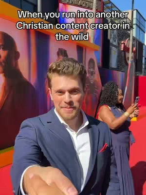 A man in a blue suit smiles at the camera, holding out his hand. Behind him, there's a red carpet and a backdrop featuring movie posters. A woman with dark hair stands to the right, looking at her phone. The image has a vibrant, event-like atmosphere.