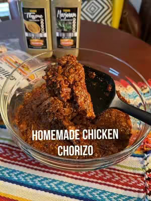 A close-up shot of a clear glass bowl filled with homemade chicken chorizo. The chorizo is a deep reddish-brown color, with a slightly textured appearance, suggesting it is finely ground. A black spoon rests in the bowl, partially submerged in the chorizo for serving. In the background, out of focus, are two bottles of spice jars, and parts of a colorful tablecloth. The text "HOMEMADE CHICKEN CHORIZO" is overlaid on the image in a white font.
