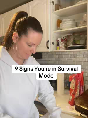 The cover image features a woman in a white top, with a ponytail, standing in front of a kitchen setting, where the text overlay appears below her, with text reading '9 Signs You're in Survival Mode'. The background includes kitchen cabinets, a partially open cabinet to her side, and some other kitchen elements on the right.