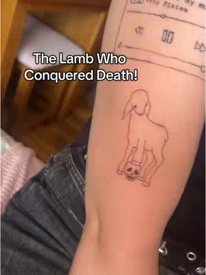 A close-up shot shows a person's arm with a tattoo of a lamb holding a skull. Above the tattoo is a digital music player bar with the name "The Pixies." The arm is positioned with the tattoo visible, the person is wearing jeans and positioned in front of a wooden background.