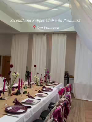 An overhead shot of a dinner party table setting. The table is long, with a neutral-colored tablecloth. The setting includes elegant plates, glassware, and vibrant purple napkins and decorations. The background suggests a well-lit room with white curtains draped along the wall.