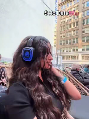 A woman with long, wavy, brown hair is wearing headphones and looking to her right. She is sitting outside with a building in the background. Text overlayed on the image says 'Solo Date'.