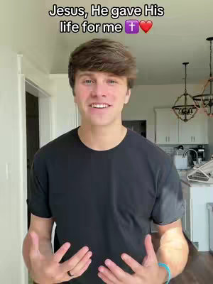 A young man in a black t-shirt stands indoors, smiling and gesturing with his hands. Overlay text at the top reads 'Jesus, He gave His life for me' with a cross and heart emoji.