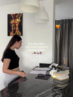 A medium shot of a woman standing in front of a desk, likely in a home environment. She is seen from the side, looking down toward items on the desk. The setting appears organized, with visible objects including books and papers next to a computer. A piece of abstract art hangs on the wall behind the woman, with a white ceiling and a curtain partially visible. The overall setting suggests a dedicated workspace or study area.