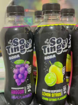 An image of several So Tinge soda bottles, each featuring a black label with white text and colorful fruit illustrations. The bottles have black caps and are arranged closely together.