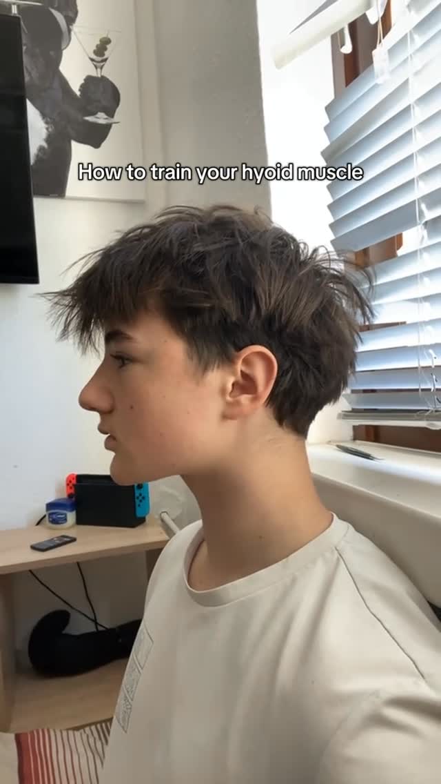 A young man is seen in profile, looking towards the right. The image has text overlayed. The background includes a window with blinds and a partial view of a room with some objects on a small table. The lighting appears natural and the composition focuses on demonstrating the hyoid muscle.
