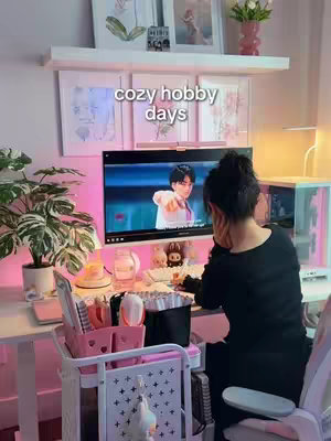 A person sits at a desk with a computer, a monitor displaying a person in a white shirt. A shelf with framed pictures and a vase of flowers sets above the monitor. A cart with supplies sits to the left, and a pink wall is lit with pink lights to the left.