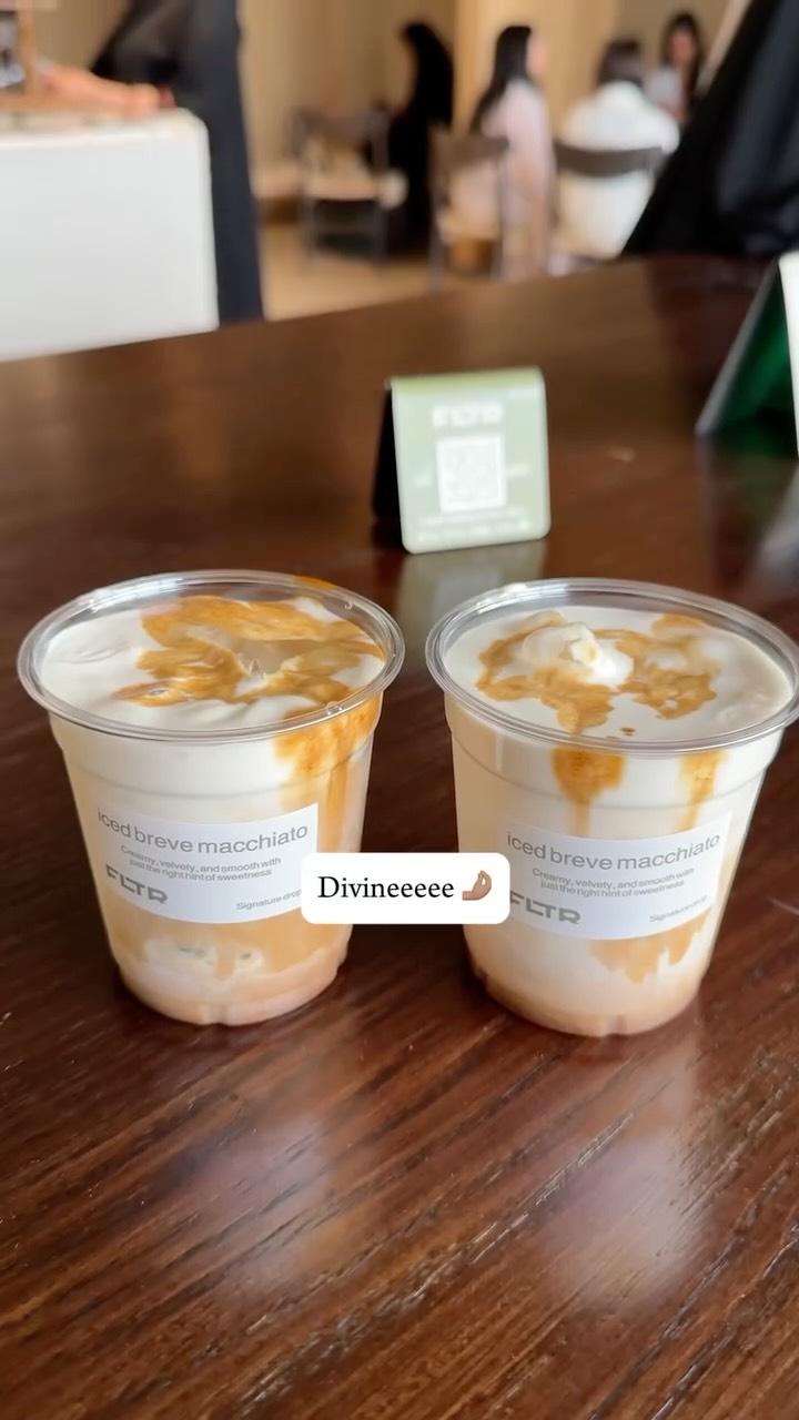 Two clear plastic cups each filled with a creamy coffee beverage, topped with frothed white milk and a swirl of caramel-colored drizzle. The cups are on a dark wooden table. There's also a green sign with a QR code nearby.