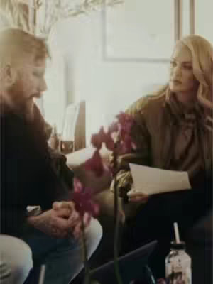 The image showcases a man and a woman seated indoors, potentially in a living room. The man, on the left, has a beard while the woman has blonde hair and is holding a paper in her hands. Between them, a vase of flowers with purple flowers stands prominently, adding depth to the scene. The warm lighting and the casual setting give a vibe of intimacy and casual conversation. The background shows interior elements such as walls and what appears to be a window or door.