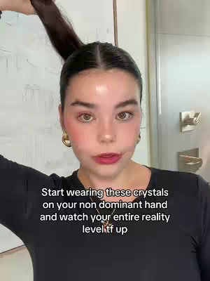 A woman with fair skin and dark hair is fixing her hair against a blurred background. The woman has prominent blush and a dark lip color, accessorized with gold hoop earrings and a necklace. Overlayed text is placed at the bottom of the image in a clear font.