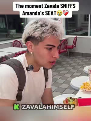 A person with short, styled, blonde hair wearing a white t-shirt and a backpack is sitting at a table in a restaurant. Text overlays the image at the top. The person is looking down and to the left, and a microphone is attached to the shirt.