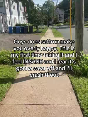 A selfie-style video taken from a low angle while the creator walks along a sidewalk. The sidewalk runs horizontally across the lower part of the frame. On the left are older brick buildings, and the right side features green grass and a street with a pole in the foreground. The sky is visible at the top of the frame. Text overlay in the image suggests a conversation about the creator's experience with saffron.