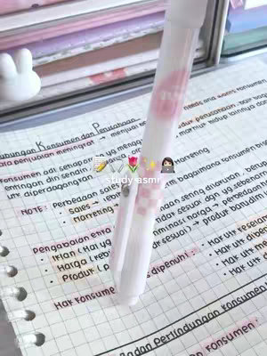 A close-up shot of a notebook page filled with handwritten notes in Indonesian. A white highlighter pen with pink accents is positioned diagonally, partially obscuring the text and drawing focus. The background includes other stacked notebooks and a small, white decorative bunny figure, suggesting a study environment.