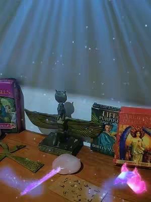 An indoor scene featuring a wooden table with multiple objects. There are books, figurines, a crystal, and cards, with some objects partially out of view. The lighting is soft, with light rays coming from above, suggesting sunlight or a spotlight, creating a celestial or spiritual atmosphere. There are sparkling effects added to the scene.