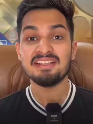 A man with a short beard and short, dark hair is centered in the frame, looking directly at the camera with a slight smile. He wears a black shirt with white trim around the collar and has a microphone clipped to it. The background is blurred, likely a room with a brown chair.