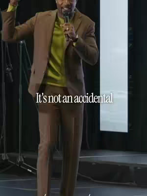 A man in a brown suit and a green shirt is speaking into a microphone on a stage. Text that reads "It's not an accidental" is overlaid on the image.