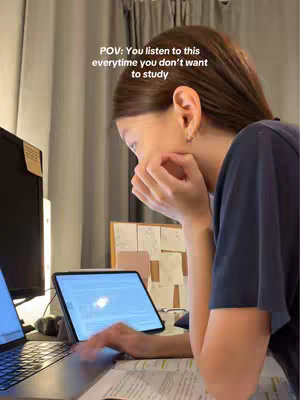 A woman with dark hair sits at a desk, resting her head in her hand while looking at a tablet screen in front of her. A laptop is to her left and a computer monitor is behind her. The scene suggests a study or work environment at home.