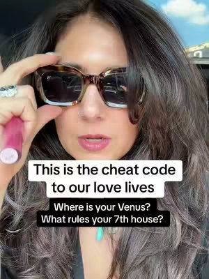 A woman with long, wavy dark hair is shown from the chest up against an out-of-focus background. She is wearing large, square, tortoise-shell sunglasses and a black top. Her left hand is raised, holding a tube of pink lipstick near her face, as if she's about to apply it. On her left hand, a ring.