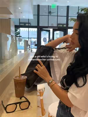 A woman is standing at a coffee shop counter with a cup of iced coffee and sunglasses in front of her. The woman is wearing a white blouse with dark jeans.