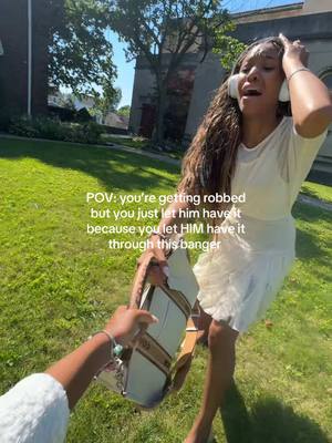 A woman in a white dress and headphones is outside on a sunny day. She is holding up her hands towards her head, seemingly in distress. She is on a well-kept green lawn with some greenery in the background, and a building is visible in the distance.  A hand holding a purse is reaching out to her, which suggests someone trying to rob her. The hook text is overlaid with a white font with a black outline.