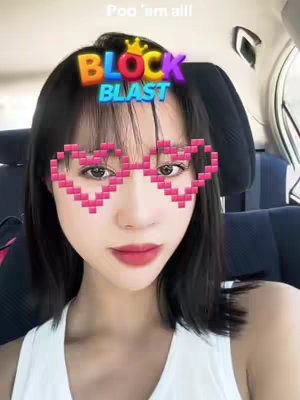 A selfie-style photo shows a fair-skinned woman with dark hair and a white tank top. Overlaid on her face are digital heart-shaped glasses made of pink blocks. Above her head is a Block Blast graphic. The background suggests she is in a car.