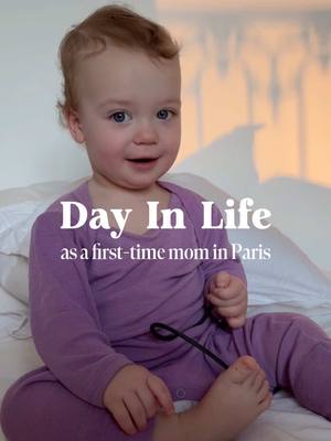 A cover photo featuring a baby sitting up on a bed. The baby is wearing a purple onesie and has light brown hair. Text is overlaid in a white font that says 'Day In Life as a first-time mom in Paris'. The background has soft lighting and the baby is smiling.