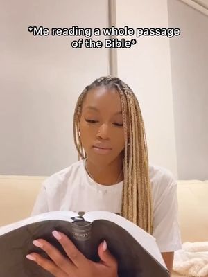 A woman with long blonde braids is seated indoors, holding a book open and looking down at it pensively. She is wearing a white t-shirt, and the image is well-lit, indicating a potentially casual, indoor setting. The background is blurred, drawing focus to her and the book.