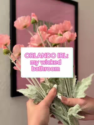 A close-up shot of a bouquet of pink flowers being held by a person with light skin. The background features a framed pink image. Overlayed on top is a white rectangle with text.
