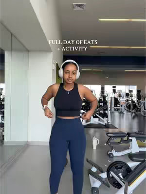 A woman is standing in the gym wearing a black tank top, blue leggings, and white headphones. Text
"FULL DAY OF EATS + ACTIVITY" is overlayed on the image in white font at the top of the image.