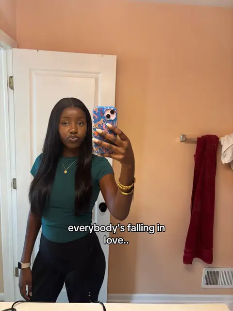 A woman is taking a mirror selfie in front of a bathroom mirror. She's holding a phone in front of her while looking towards the camera with a slight pout. She wears a green top and black leggings, gold bracelets and a watch. The bathroom has a salmon-colored wall and a white door.