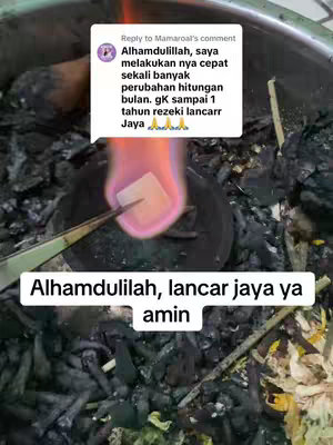The image shows a close-up view of a small, dark container filled with charred material, possibly charcoal, with a bright flame rising from the center, illuminating a small square object held in tongs. The background gives an impression of being outdoors or in a dimly lit space. A text overlay at the bottom reads a prayer-like statement in Indonesian.