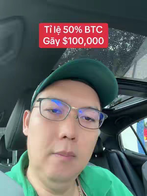 A man wearing glasses and a green cap is filming himself from inside a car. On the top left corner is a red rectangular overlay with white text announcing, "Tỉ lệ 50% BTC Gãy $100,000". The man has a serious expression.
