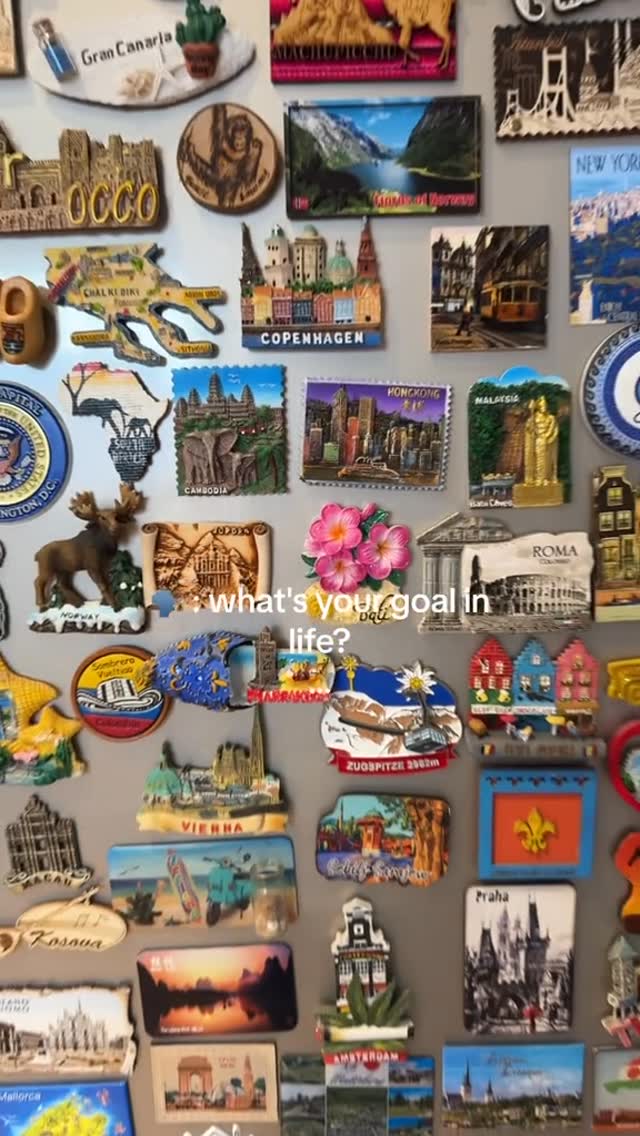 A close-up shot showcases a multitude of souvenir magnets arranged on a neutral, possibly metallic surface. The magnets depict various landmarks, cities, and cultural symbols from different locations around the world. A text overlay asks "what's your goal in life?"