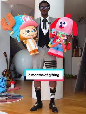 The cover image shows a man in a stylish outfit and holding stuffed toy characters in an interior setting. The man, of African descent, is wearing a gray suit jacket over a black vest, light shorts and knee-high socks with black loafers. He wears glasses and his arms are extended, supporting the two stuffed animals: a cartoon character with a blue head and another cartoon character with a pink hat and a red dog. A text bar at the bottom reads "3 months of gifting"