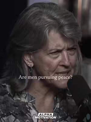 A close-up shot of a woman with gray hair and a serious expression. She is wearing a patterned top and is positioned in front of a dark background and a microphone