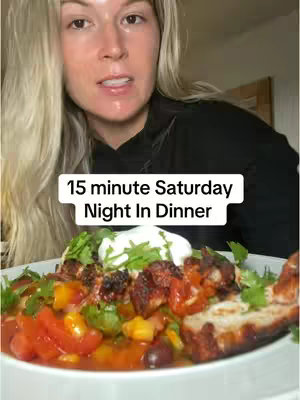 A woman with long blonde hair is smiling and looking at the camera, she appears to be holding a plate of food. Overlayed, there's text that reads "15-minute Saturday Night In Dinner". The food bowl is white, the plate is filled with a variety of ingredients bathed in a sauce, with a dollop of white cream on top and cilantro as garnish.