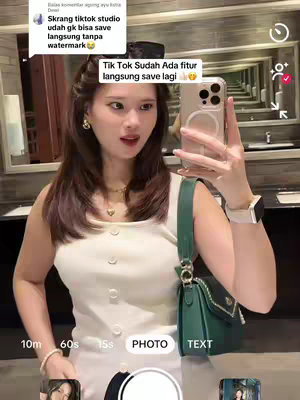 A selfie-style image featuring a young woman in a well-lit bathroom. She is holding a phone up to take a picture, displaying the screen in a selfie pose. Behind her, the bathroom features a modern design with mirrors and reflective surfaces. The woman wears a white top with button detail and a green purse, suggesting a casual, fashionable style. The overall composition is bright and clean.