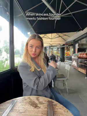 A woman sits at a table in an outdoor cafe setting, looking toward the camera with a slight smirk. She is wearing a gray blazer and blue jeans. Text is overlaid on the image.