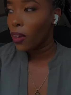 A close-up shot of a woman with dark skin looking slightly off-camera. She has her hair styled in braids, with an earbud in one ear. Her makeup includes eye shadow, mascara, and dark lip color. She wears a gray shirt and a gold pendant necklace. The background is softly blurred, suggesting an indoor setting.